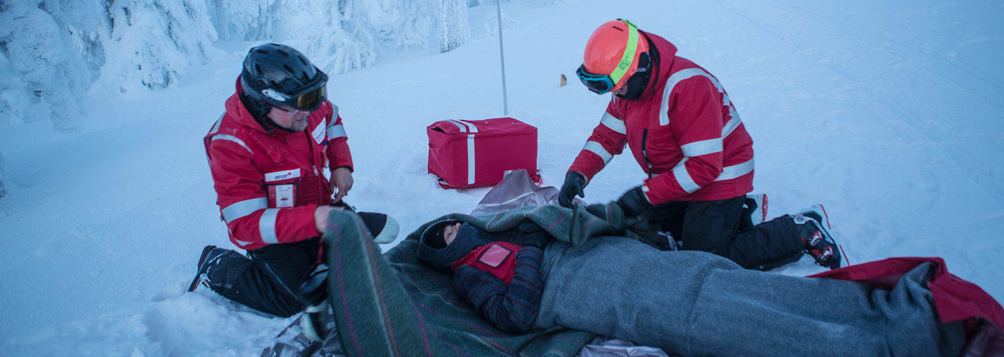 Kaksi Punaisen Ristin rinnepäivystäjää käärii huopaa lumella makaavan henkilön ympärille laskettelurinteen reunassa.