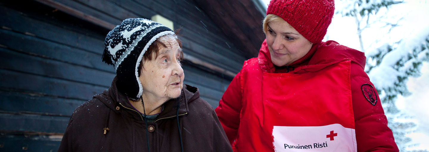 Kaksi naista seisoo rinnakkain talvisessa maisemassa. Nuorempi, Punaisen Ristin vapaaehtoisliiviin pukeutunut nainen katsoo lempeästi iäkkäämpää naista.