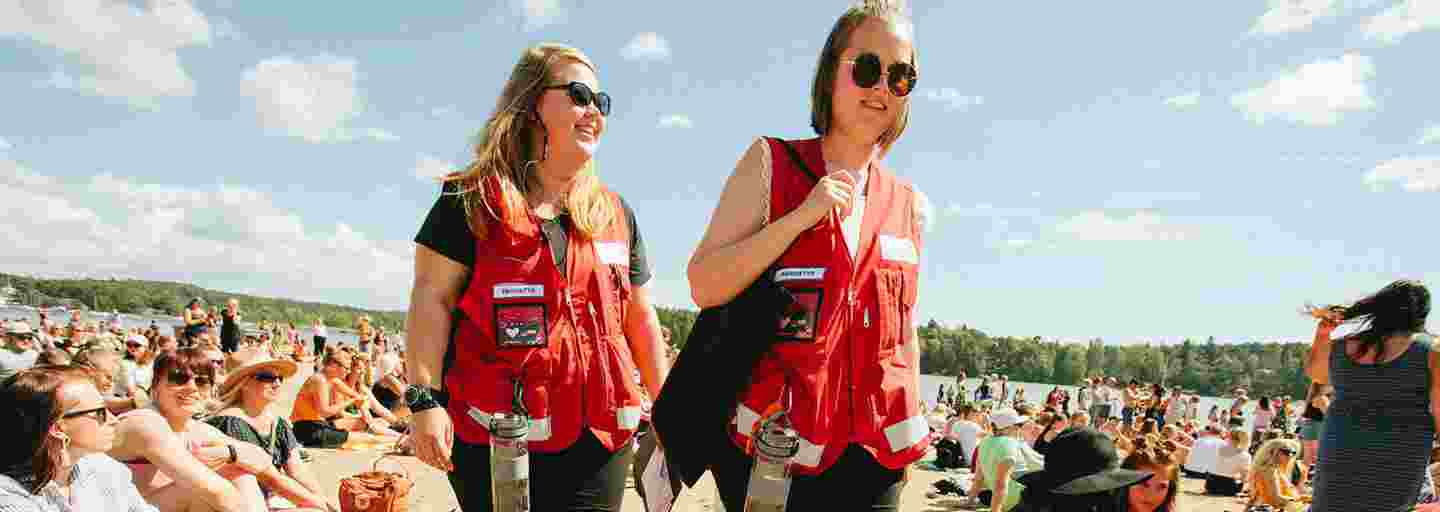 Kaksi nuorta naista Punaisen Ristin vapaaehtoisliiveissä suorittaa ensiapupäivystystä aurinkoisessa Ruisrockissa.