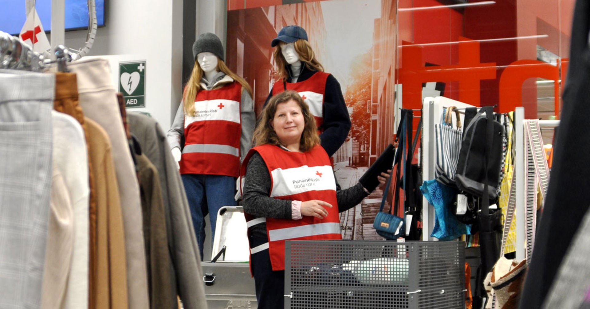 Punaisen Ristin Kontti-kierrätystavaratalossa henkilö esittelee myynnissä olevia laukkuja. Kuvassa näkyy vaaterekkejä, laukkuja sekä taustalla kaksi Punaisen Ristin liiveihin puettua mallinukkea. Punaisen Ristin tunnus näkyy useissa kohdissa myymälää.