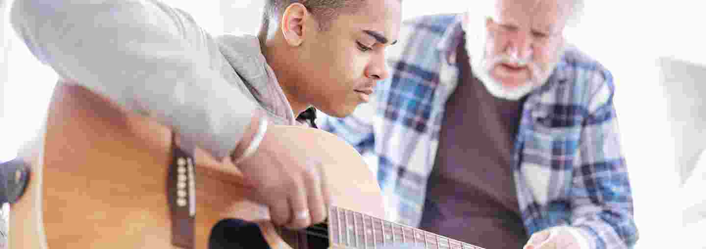 Nuori mies opettelee kitaransoittoa vanhemman miehen avustuksella.
