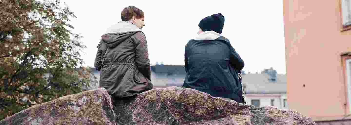 Kaksi nuorta aikuista keskustelee syksyisessä maisemassa kalliolla istuen.