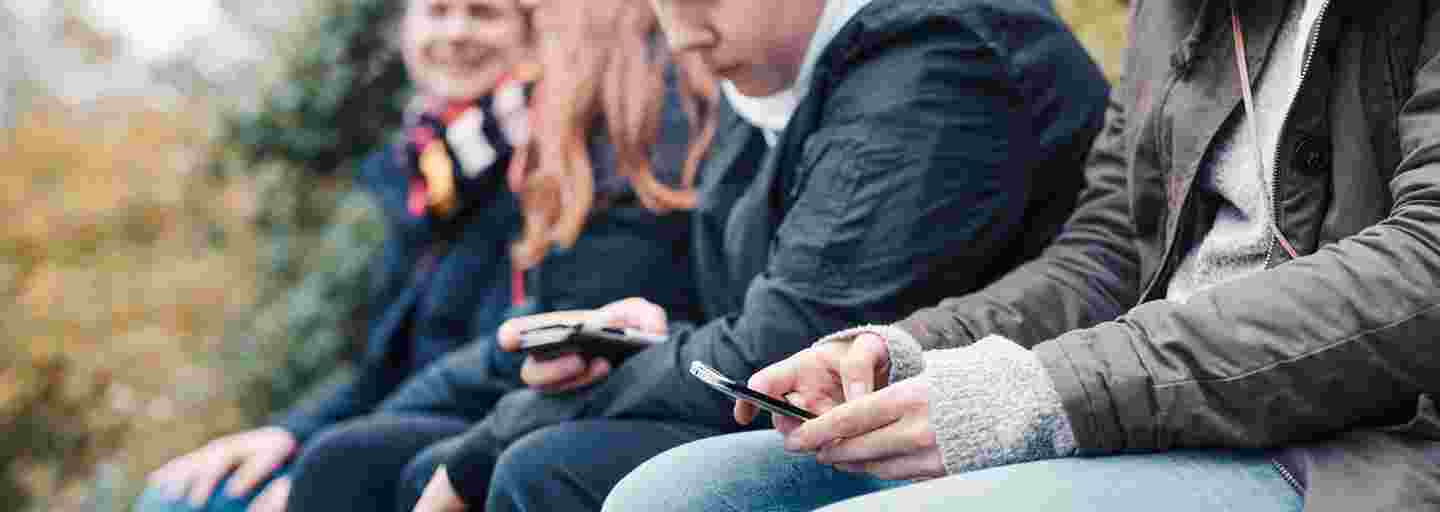Neljä nuorta istuu rivissä ulkona älypuhelimet käsissään.