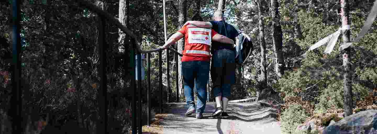 Mies tukeutuu Punaisen Ristin vapaaehtoiseen kävellessään ylämäkeä puiden reunustamalla tiellä.