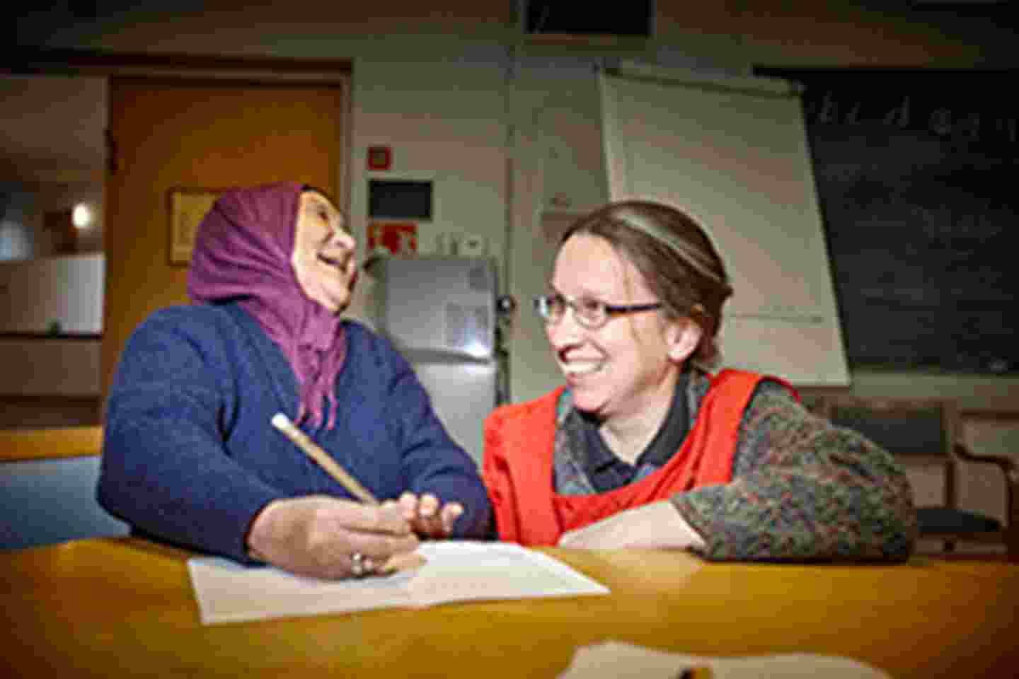 Naurava iäkäs maahanmuuttajanainen opiskelee Punaisen Ristin vapaaehtoisen opastuksella.