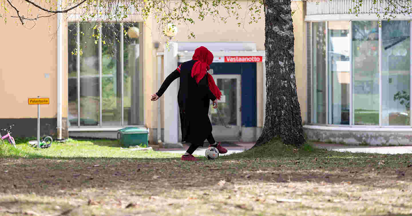 Huivipäinen nainen pelaa jalkapalloa vastaanottokeskuksen edustalla.