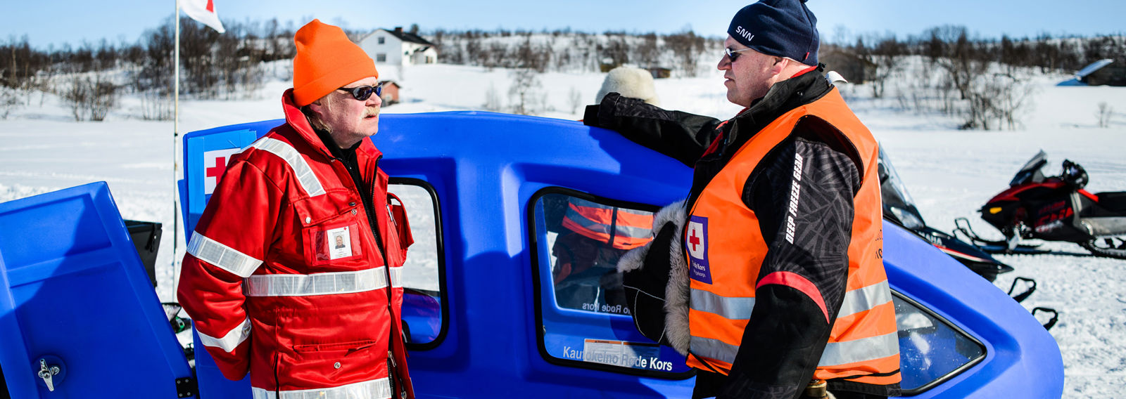 Kaksi Punaisen Ristin varusteisiin pukeutunutta miestä talvimaisemassa moottorikelkkojen vieressä.