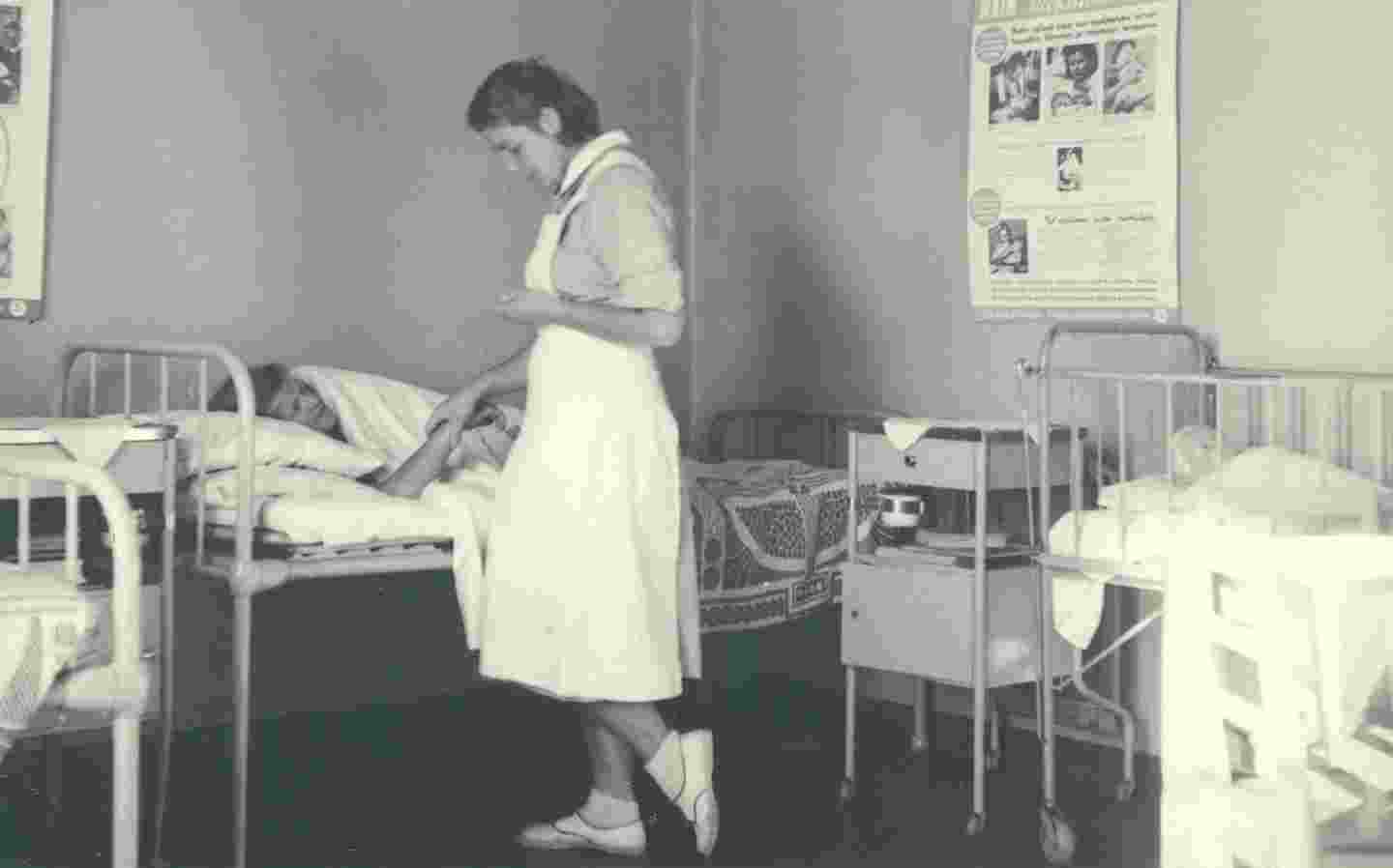 Nuori nainen Punaisen Ristin sairaanhoitaja-asussa hoitaa vuoteella makaavaa potilasta.