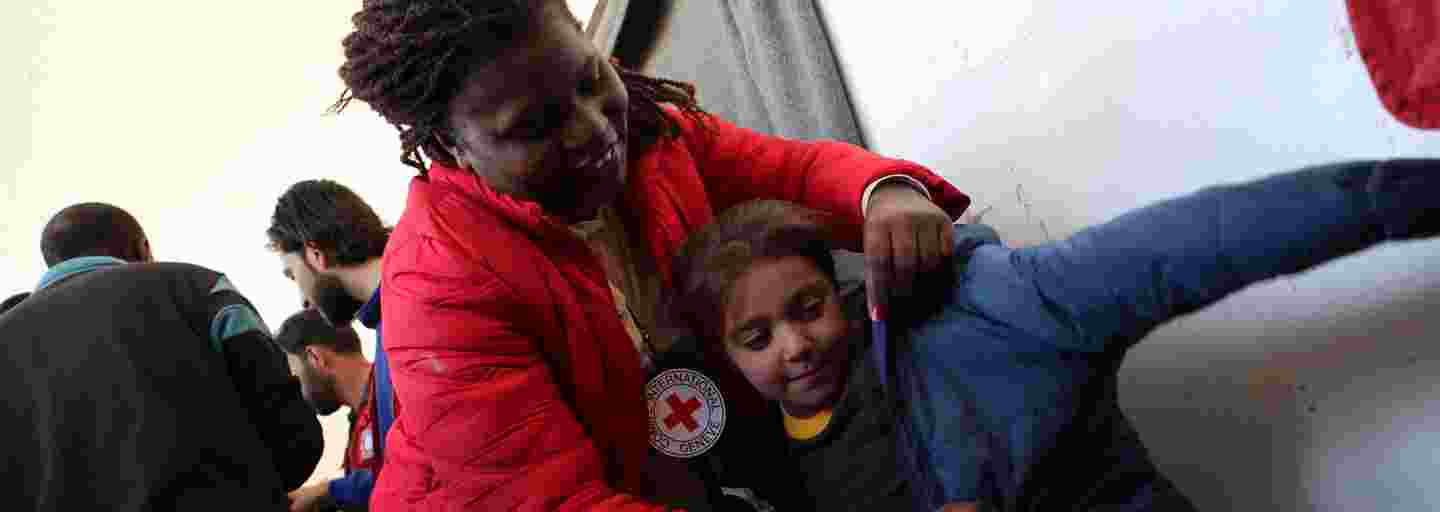 Hymyilevä Punaisen Ristin varusteisiin pukeutunut nainen pukee nuorelle tytölle takkia.