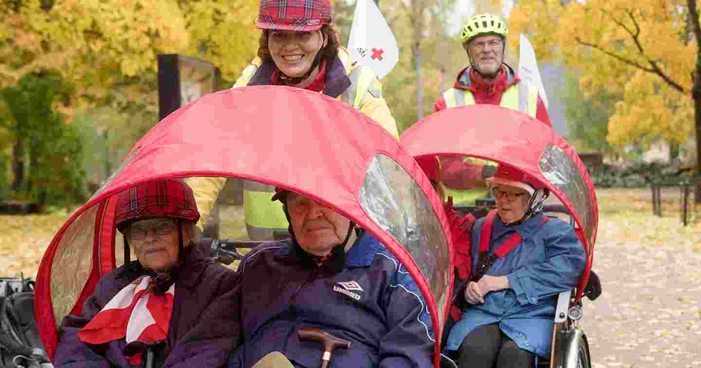 Kaksi Punaisen Ristin vapaaehtoista ulkoiluttaa ikäihmisiä riksapyörillä.