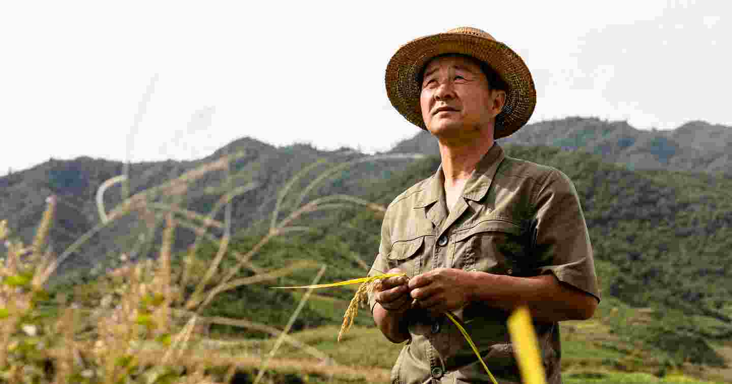 Aasialainen mies seisoo pellolla vuorten keskellä käsissään viljelyskasveja.