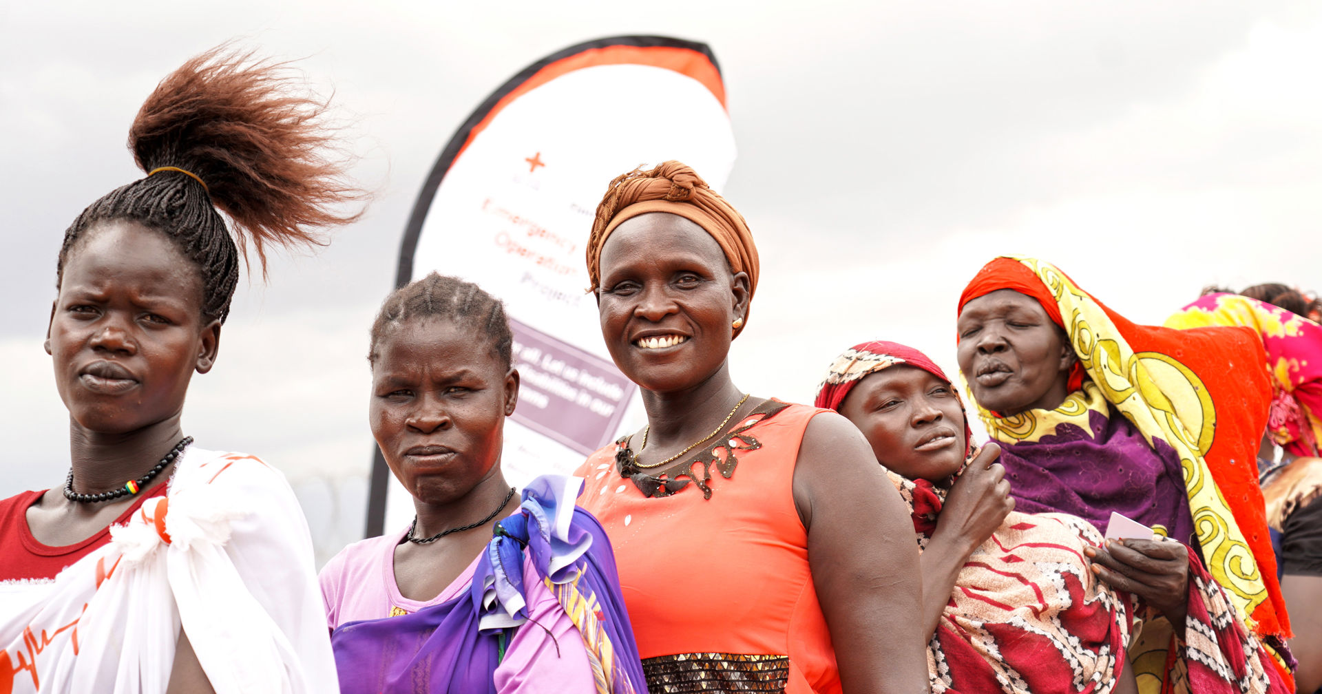 Ihmiset seisovat jonossa ulkona tuulessa takanaan banneri, jossa näkyy Punaisen Ristin logo.