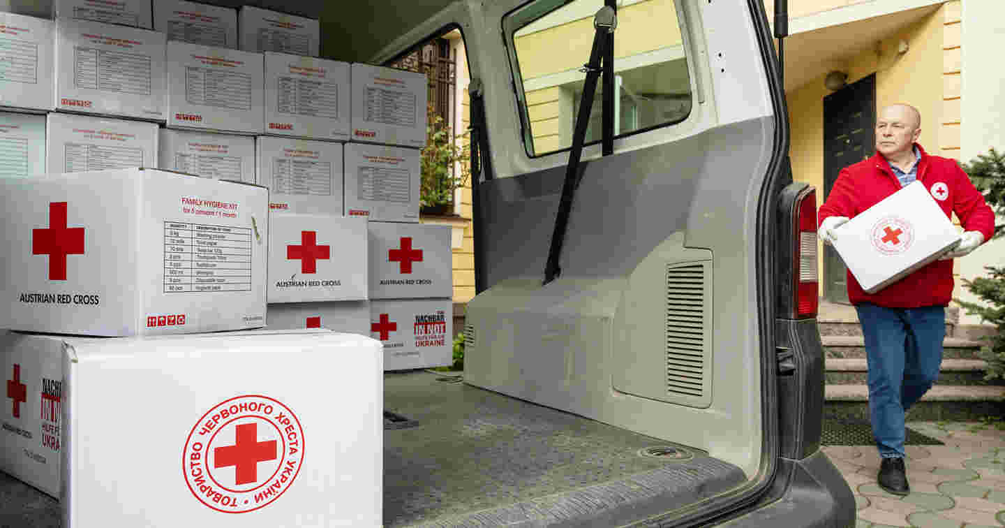 Henkilö Punaisen Ristin työasussa kantaa Punaisen Ristin avustustarvikelaatikkoa pakettiautoon, joka on puolillaan avustustarvikkeita.