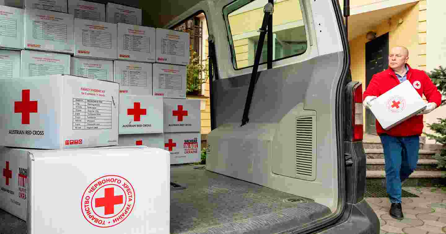 Henkilö Punaisen Ristin työasussa kantaa Punaisen Ristin avustustarvikelaatikkoa pakettiautoon, joka on puolillaan avustustarvikkeita.