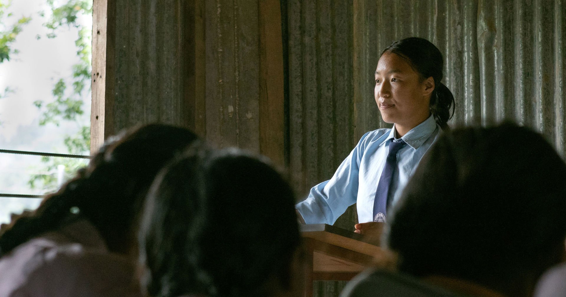 Nuori henkilö seisoo luokkahuoneessa puhujankorokkeen takana koulupuku päällään. Etualalla näkyy oppilaita kuuntelemassa, taustalla luokkahuoneen seinärakenteita ja ikkuna.