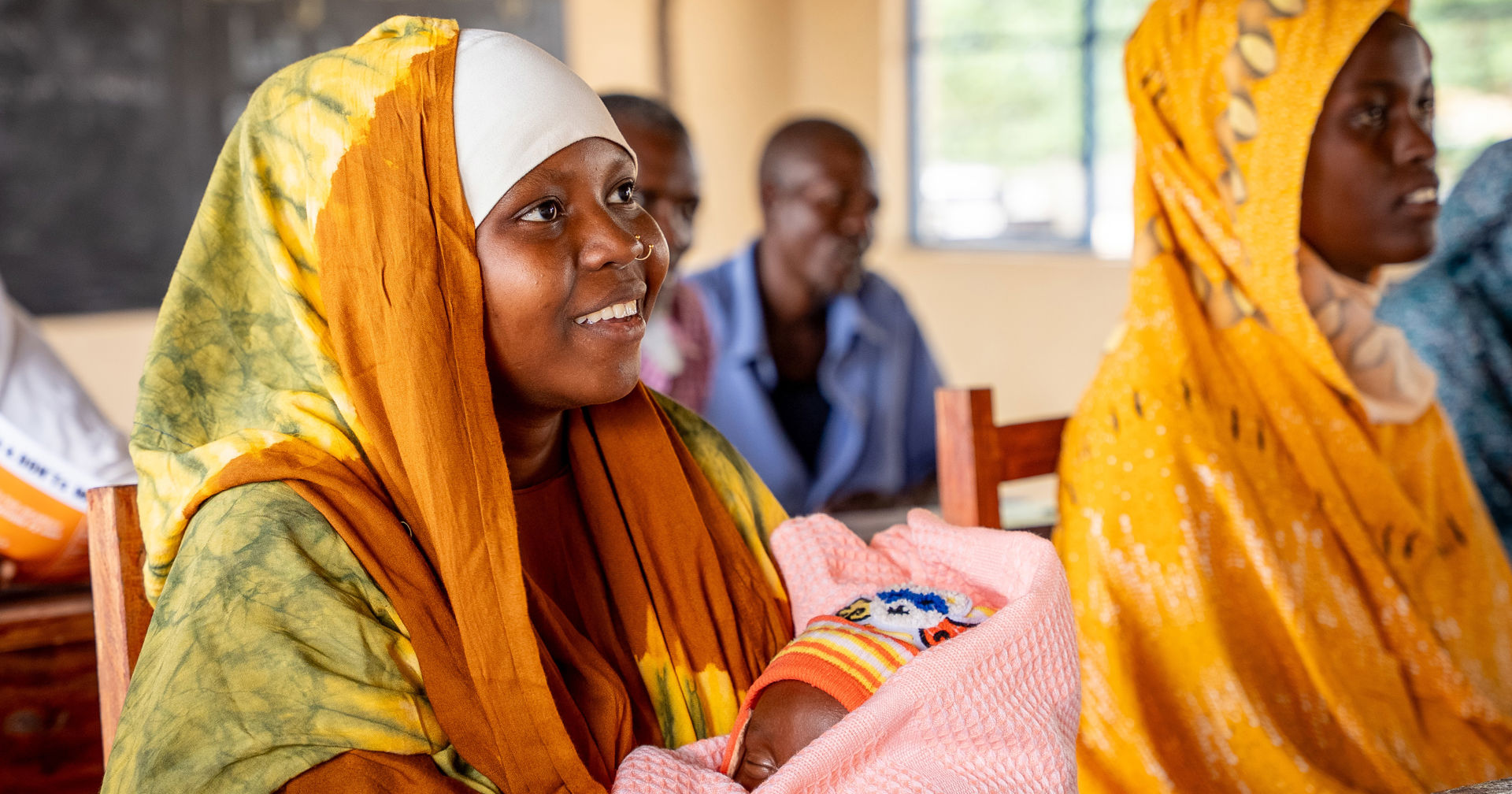 Henkilö istuu luokkahuoneessa ja pitää sylissään vauvaa, katsoo eteenpäin ja hymyilee. Taustalla näkyy muita ihmisiä istumassa.