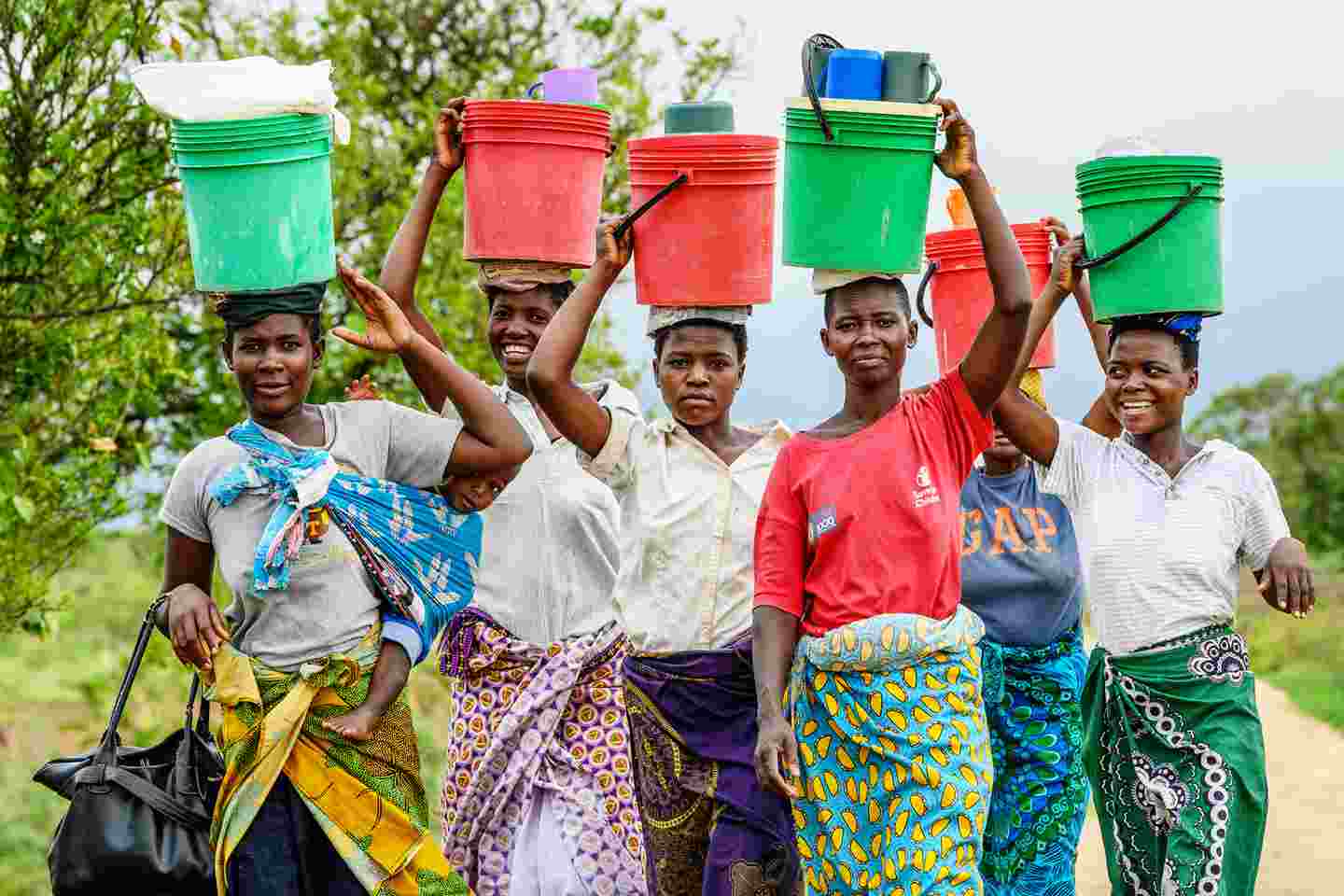 Kuusi hymyilevää naista kantaa vettä ja tarvikkeita päänsä päällä.