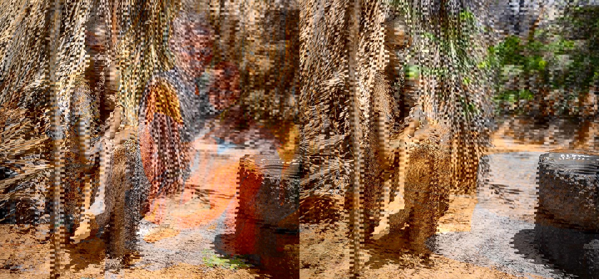 Nainen istuu lapsi sylissään takanaan savesta ja lehdistä rakennettu rakennus.