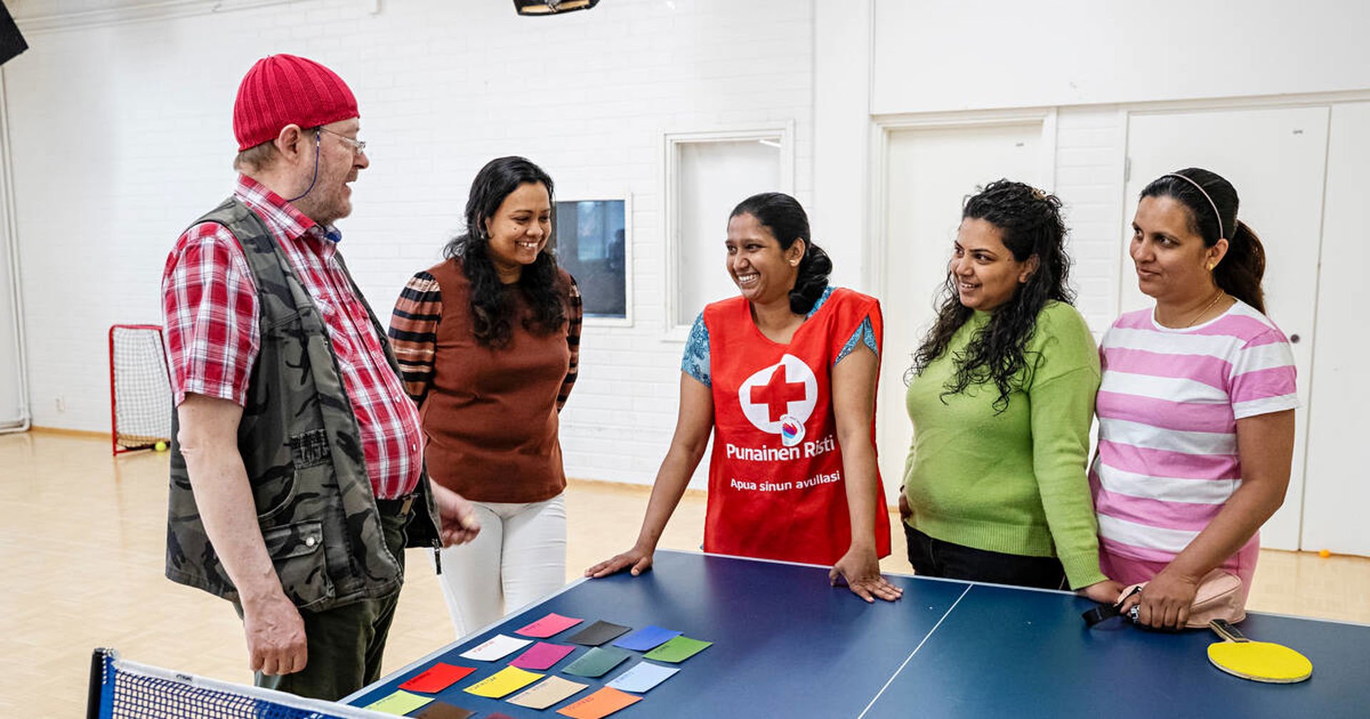 Joukko ihmisiä pelipöydän äärellä, keskimmäisenä olevalla naisella on Punaisen Ristin vapaaehtoisliivi.