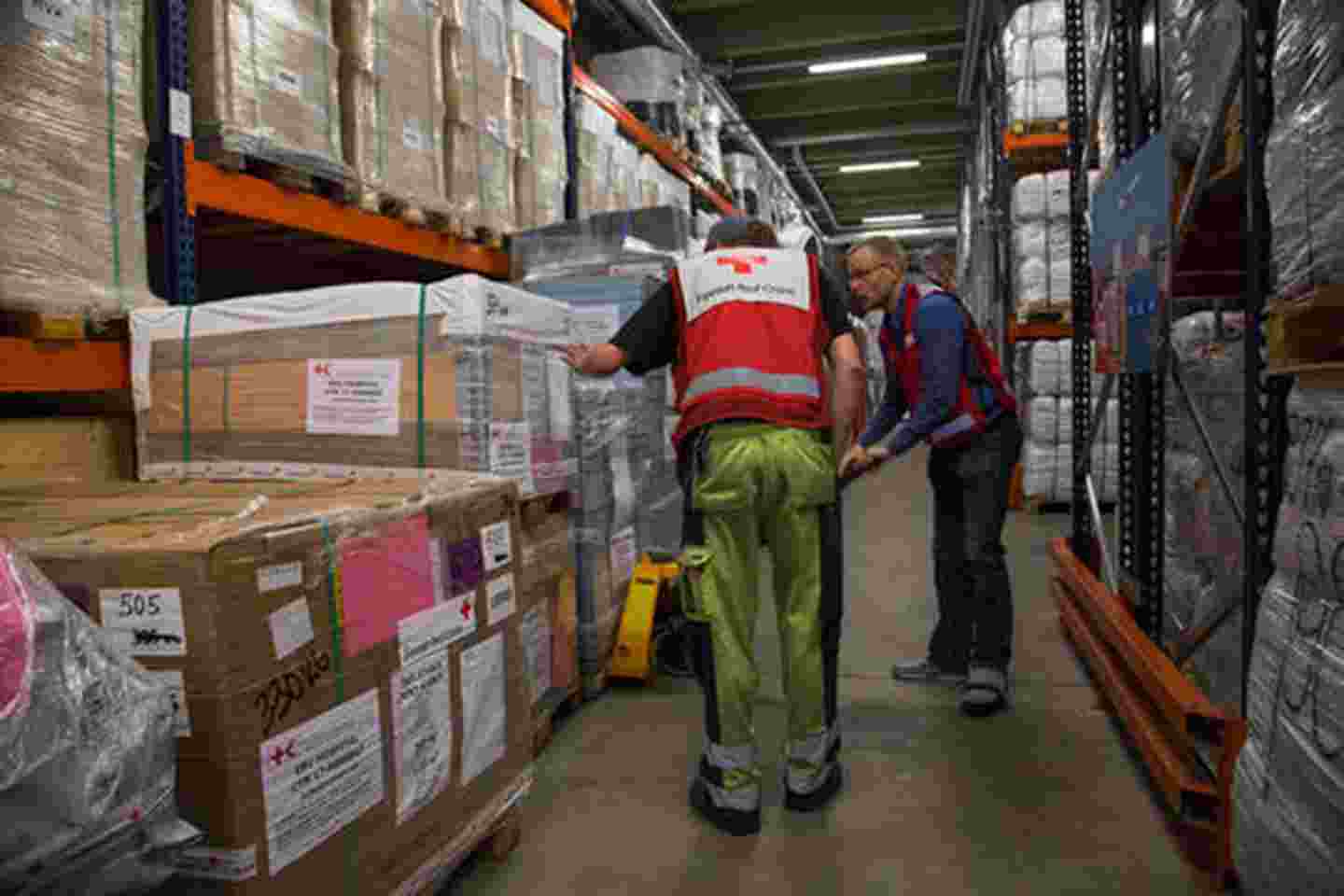 Kaksi Punaisen Ristin työntekijää noutaa avustustarvikkeita logistiikkakeskuksessa, jonka korkeat ja pitkät hyllyt ovat täynnä tarvikkeita.