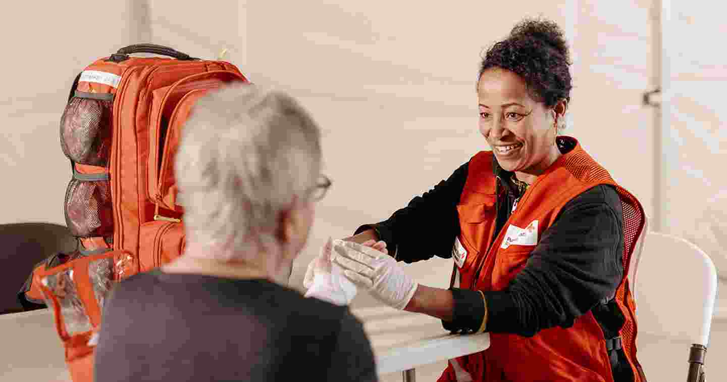 Hymyilevä henkilö Punaisen Ristin vaatteissa tekee sidosta iäkkään henkilön käteen.