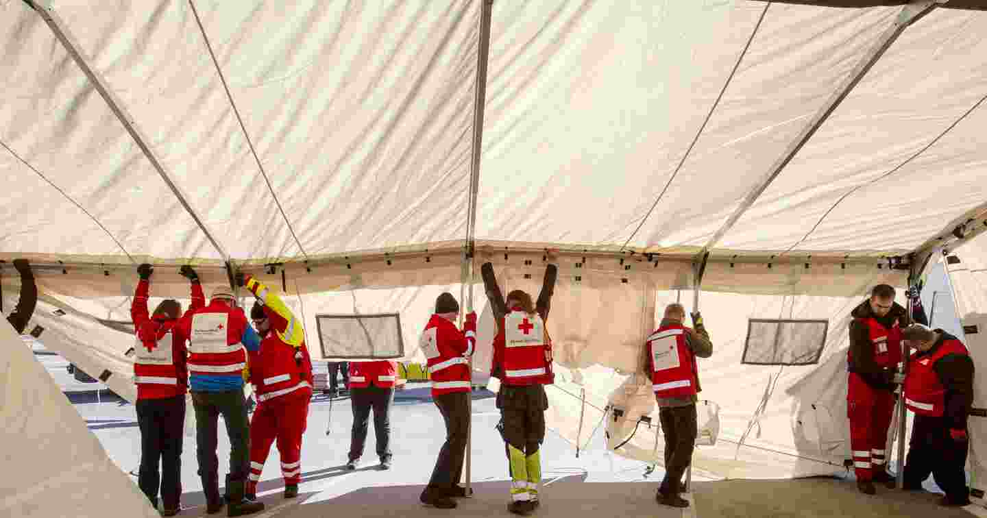 Yhdeksän henkilöä Punaisen Ristin vaatteissa pystyttää isoa telttaa.