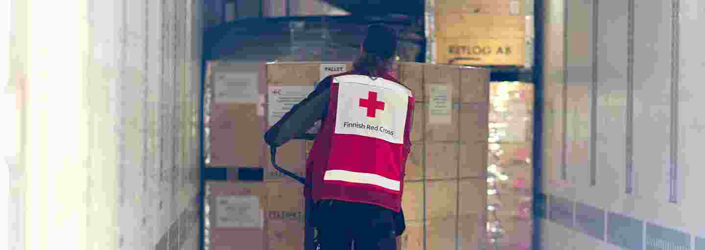 Punaisen Ristin työntekijä kuljettaa isoja avustustarvikelaatikoita kärryssä varaston käytävällä.