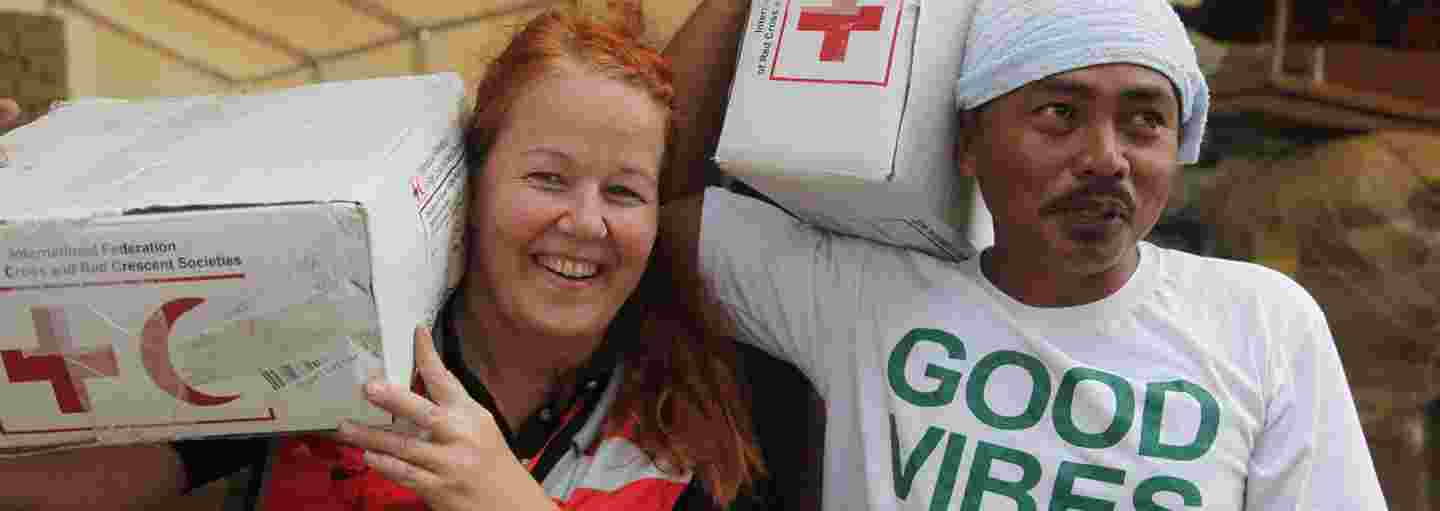 Punaisen Ristin avustustyöntekijä ja paikallinen auttaja, kantavat avustustarvikkeita hymyillen.