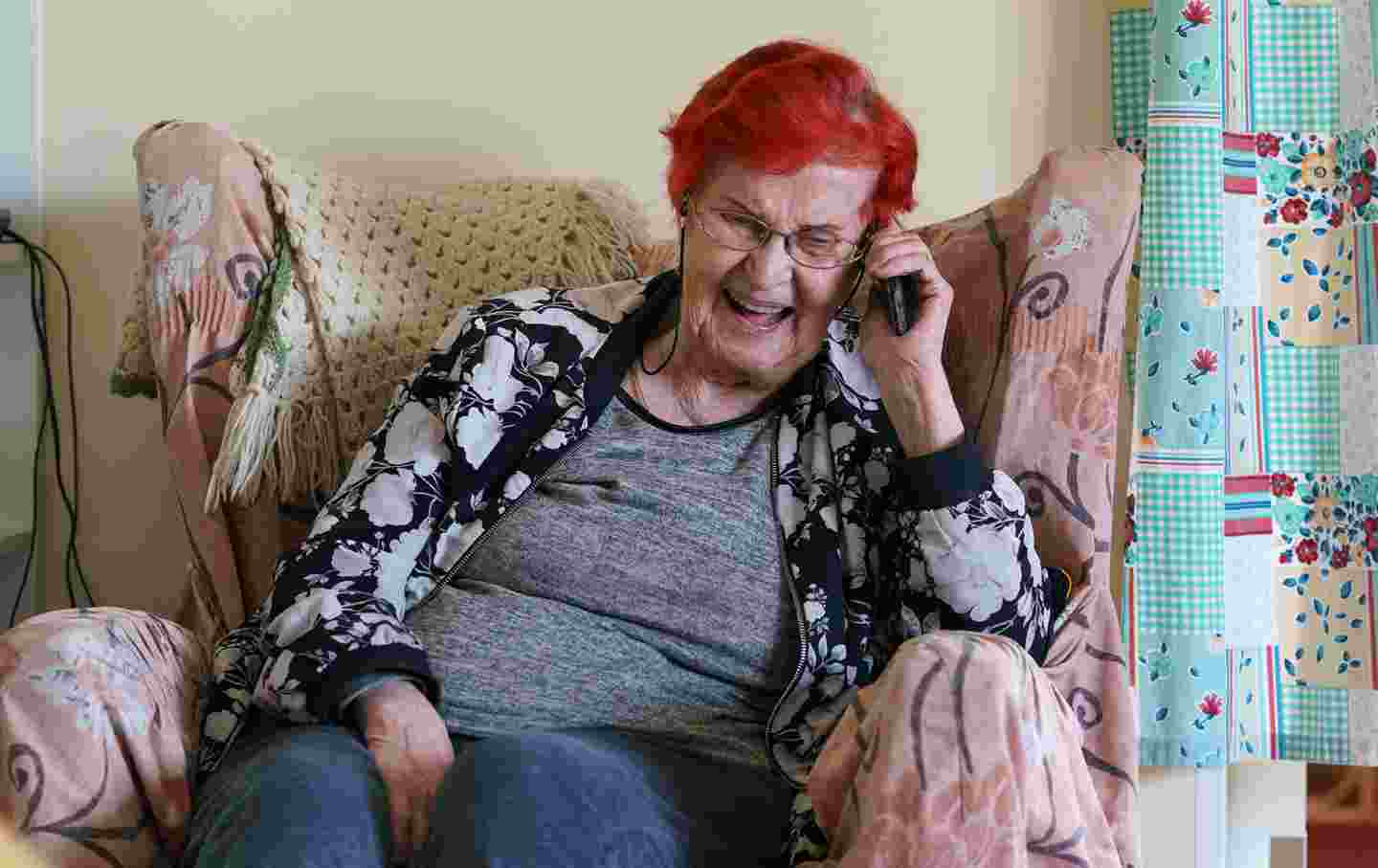 Hymyilevä iäkäs nainen puhuu puhelimeen kotisohvallaan.
