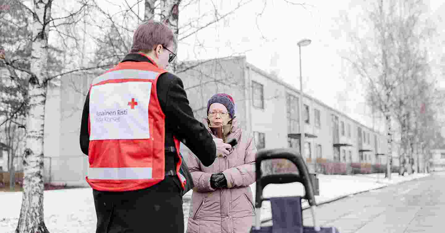 Punaisen Ristin vapaaehtoisliiveihin pukeutunut henkilö näyttää kännykältä tietoa ikääntyneelle henkilölle.