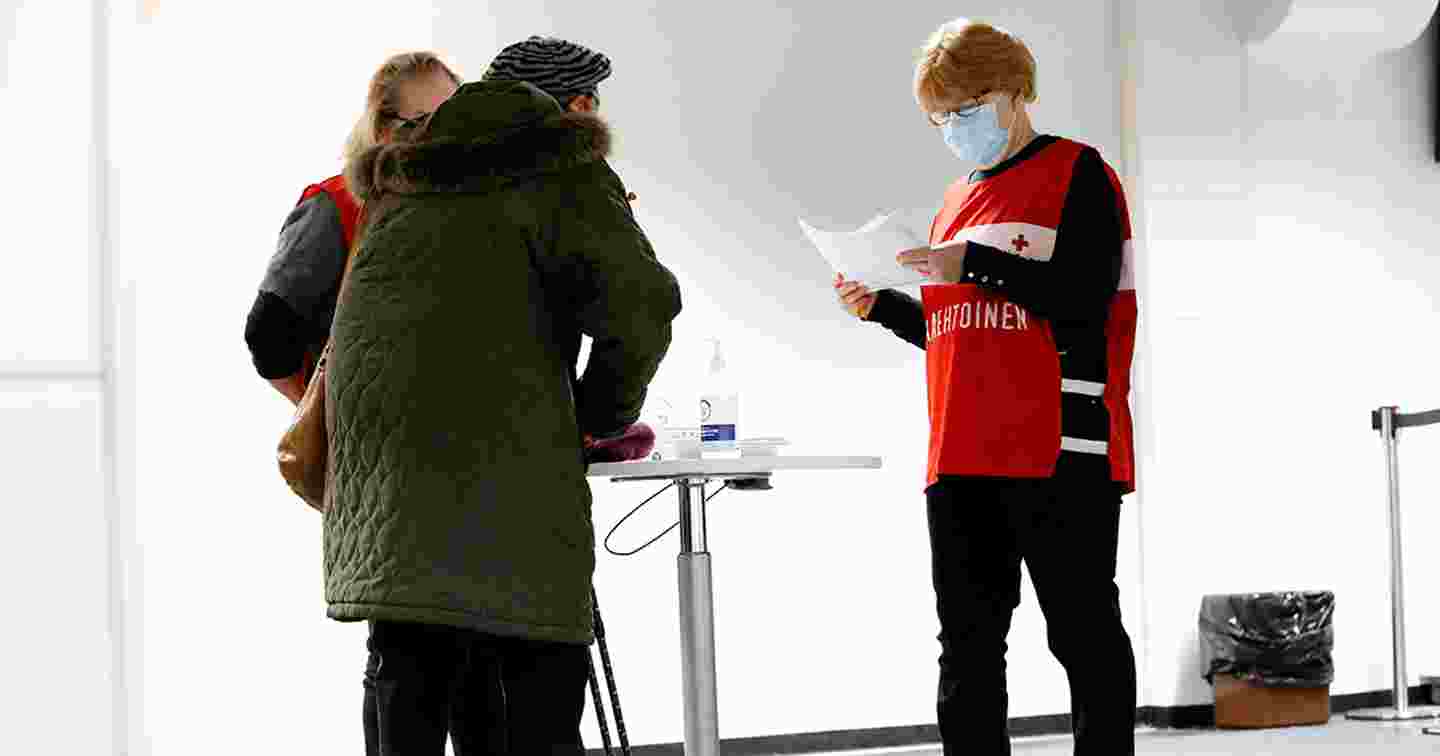Punaisen Ristin vapaaehtoisliiveihin pukeutuneet kaksi ihmistä neuvoo ikääntynyttä henkilöä tämän tullessa rokotuspisteelle.