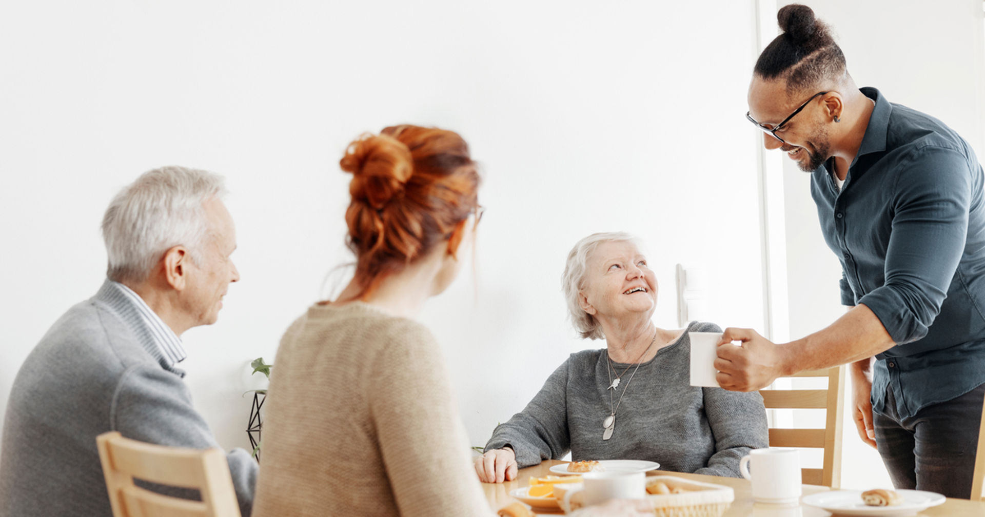 Neljä henkilöä istuu ja seisoo ruokapöydän ääressä valoisassa huoneessa, pöydällä on kuppeja, lautasia ja leipäkori, ja yksi henkilö ojentaa mukin toiselle.